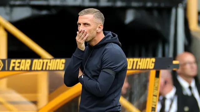 Gary O'Neil, in a black Wolves tracksuit, puts his right hand to his mouth as stands in front of a yellow dugout with the words 'fear nothing' written on the top while watching Wolves' defeat to Chelsea. 