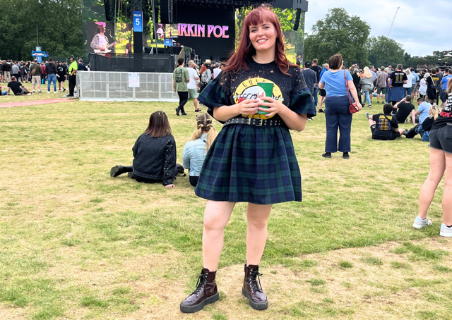 Blaize is stood in front of a stage at a festival wearing a Guns 'N' Roses band t-shirt. She is smiling. 