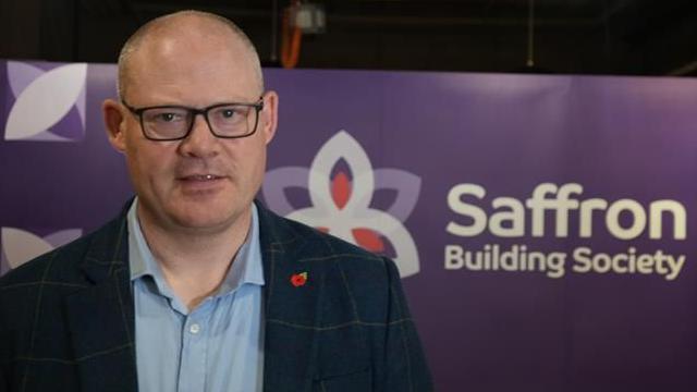 Colin Field, who is wearing a blue shirt and navy suit jacket. He is wearing glasses and looking at the camera with his mouth slightly open. He is standing in front of a purple banner that says "Saffron Building Society".