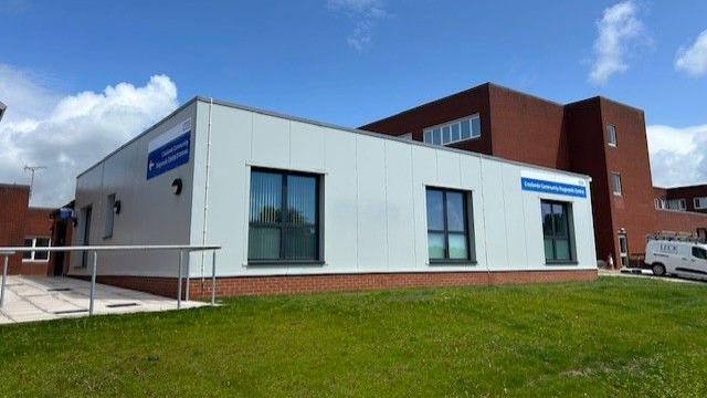 The Croslands Community Diagnostic Centre. It is a square grey building with a blue sign which states its name. Furness General Hospital can be seen behind it and a green lawn stands in front of it.