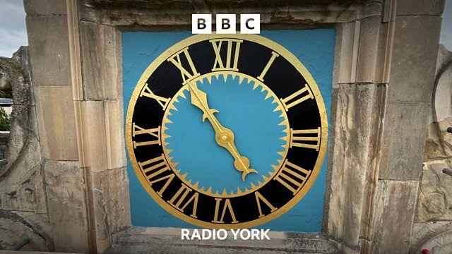 York's one-handed clock pointing to 11 on it's face of gold roman numerals on a black dial mounted on a light blue background.