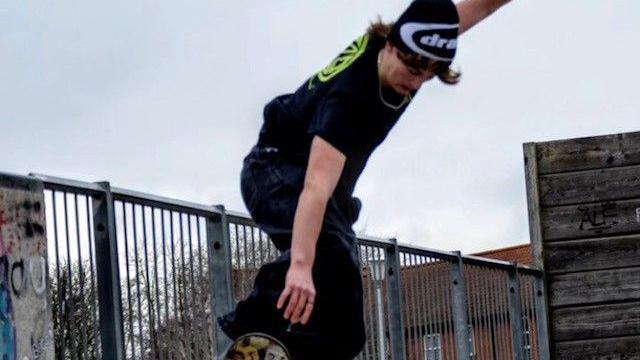 Ryan is pictured skateboarding at a skatepark. He is riding the board with one arm beside him and the other in the air. He is wearing a black and white beanie hat and a black t-shirt and trousers. 