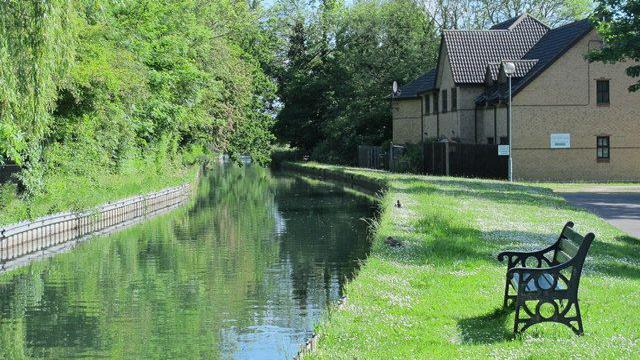 River curving round to the left with hedge on the left bank behind log roll, and grass on the right with a dark-coloured bench looking towards the river. There are new houses in the background.