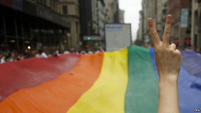 Close up of woman's hand making V for Victory sign in front of a large rainbow-coloured gay pride flat during New York Gay Pride march, 2015