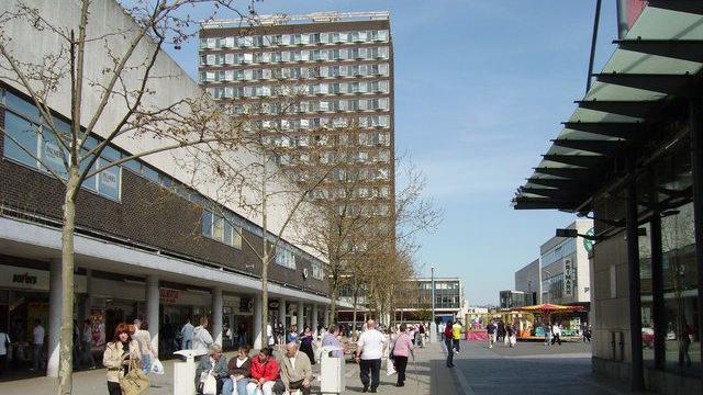 A large building with lots of windows is in the background, towering above the surrounding buildings. Large concrete buildings are in the foreground with shoppers walking or sitting on benches in a pedestrianised area.