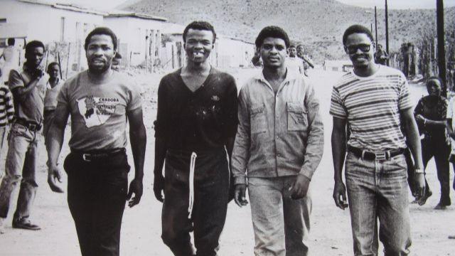 Four men walk towards the camera, smiling, in an old black-and-white photograph