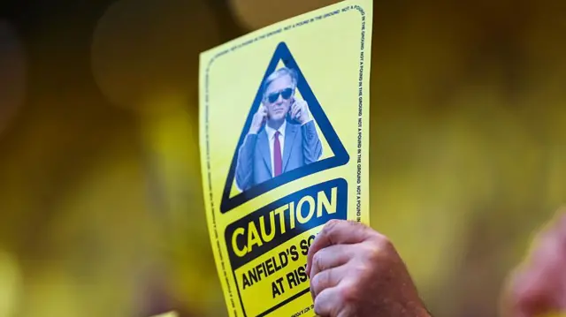 A yellow card features John W Henry on it. It reads 'Caution - Anfield's soul at risk'