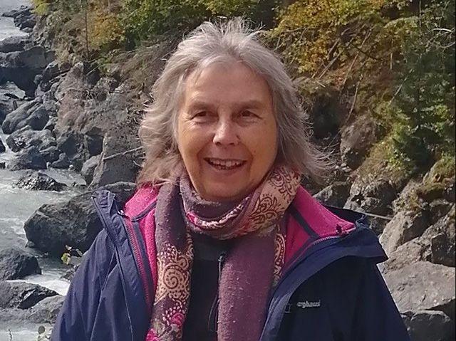 Anne Page has grey hair and a smile. She is wearing a purple patterned scarf and a purple coat. She is standing by a rocky sea shore.