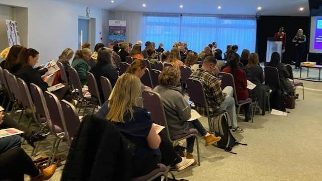 People scan QR codes on paper documents in their laps with their phones as they sit in a conference room during a conference. The attendees all appear to be adults, with eight rows of chair seen in the portion of the room photographed. Two young women can be seen leading the conference at the front of a room, with a big screen behind them.