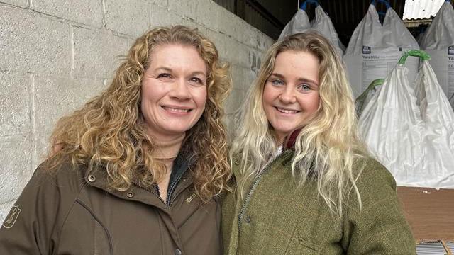 A blonde woman with curly hair in a brown coat smiling and standing next to a younger blonde woman in a green tweed coat.