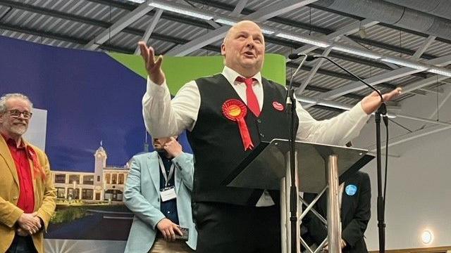 Andy Holdsworth, wearing a red rosette, giving a speech 