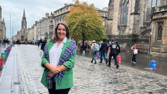 Dr Thompson is standing on the cobbles in the Royal Mile. She is wearing a white shirt and green suit jacket. She has a tartan shawl over her shoulders. She has shoulder-length brown hair and is smiling at the camera.