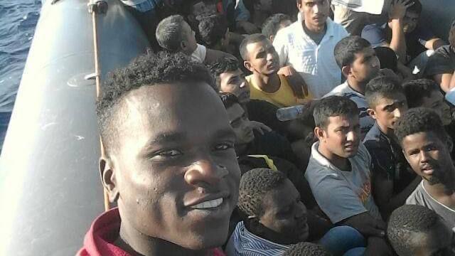 A number of young men crammed into a small boat with one person looking straight into the camera and smiling. There is a hint of blue water to the left of the picture.