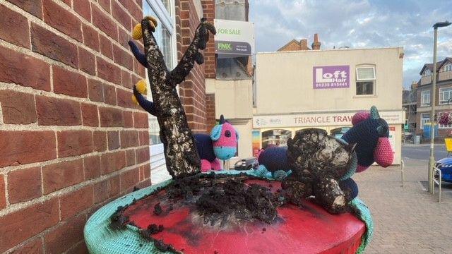 A burnt post box topper with colourful cows and a tree all black with burns. The post-box is also marked.