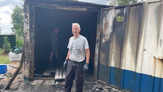 Pete Baker wearing a grey t-shirt and dark trousers, stood in front of the same burned out container as the one seen in the first picture. Another person can be seen inside the container.