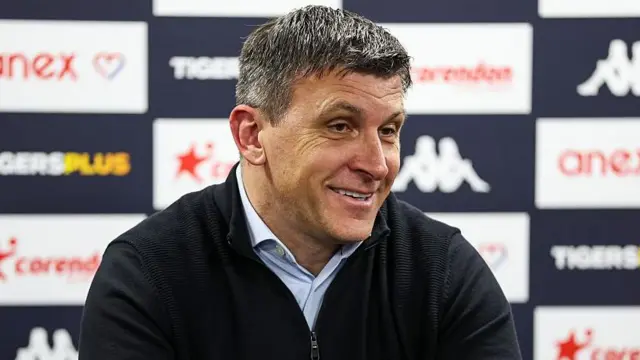 A smiling Sergej Jakirovic wearing a blue jumper and light blue shirt at a press conference in front of a sponsors' board