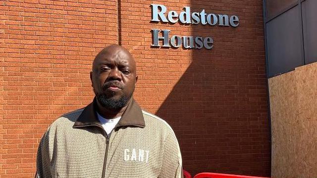 A man with short dark hair and a beard, wearing a beige zip-up jacket. He is standing in front of a brick wall with "Redstone House" in silver lettering on it. There is wooden boarding around the entrance porch, and a low red temporary fence in front of the entrance.