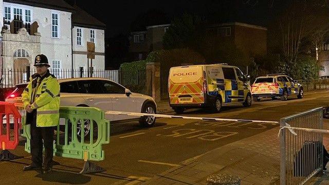 A police officer is standing at a green barrier next to police tape blocking a road, with vehicles, including a police van and car, beyond