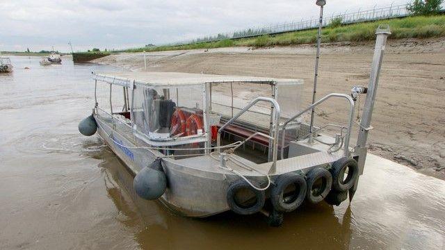 Image shows a ferry siting in the tidal waters of the River Great Ouse at King's Lynn, with buoys hanging over the side and tyres suspended at the back