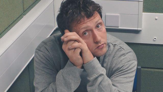 CCTV image of france in police custody. He is in a police interview room with grey brick walls. He is wearing a grey jumper and has curly brown hair. His elbows are resting on the table with his hands up against the left side of his face, He is looking directly at the camera.
