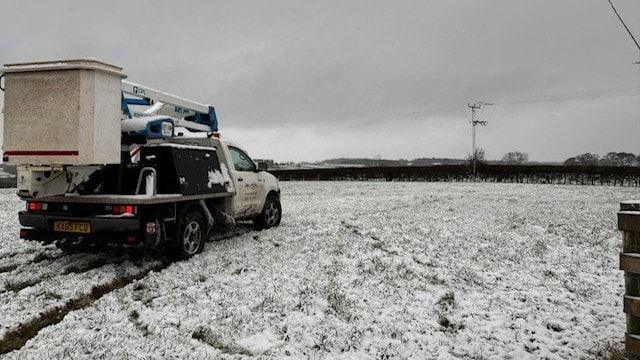 A white Electricity North West van parked in a snowy field. There is a white and blue cherry picker on the back of the vehicle. A hedgerow can be seen at the end of the field. An electricity mast stands by the hedges.