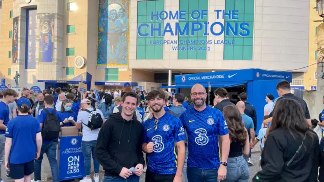 A group of three men pose for a picture in front of Stamford Bridge. A sign above them reads: 'Home of the Champions - European Champions League Winners 2021'