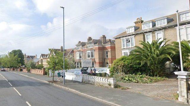 Road with large three story houses to the right and blue and white hotel sign to the left with cars in the hotel car park