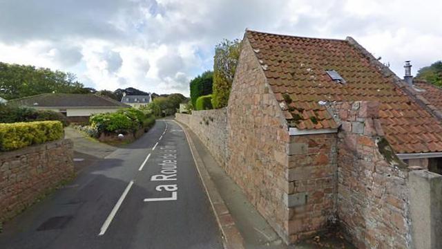 A small road with a granite building on one side and houses on the other. A small pavement runs along the right side of the road.