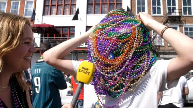 BBC reporter Katie Smith speaking to a man with his head covered with beads at a parade before Super Bowl 59