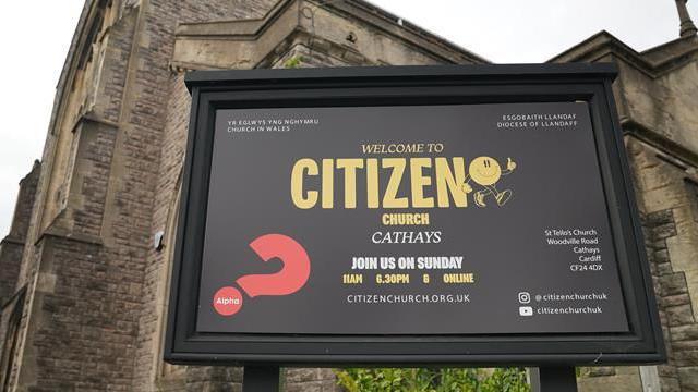 A black sign in front of a large stone church building. On the sign there is yellow writing which says 'welcome to citizen church, Cathays'