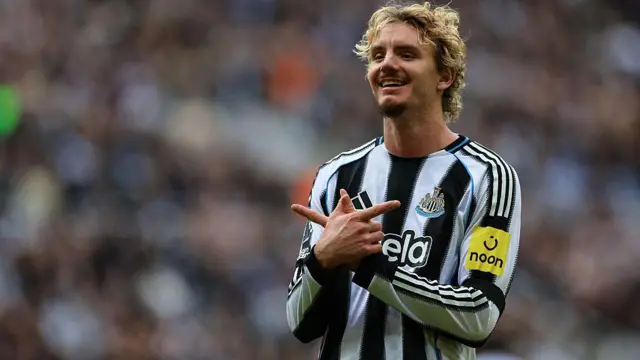 A  smiling Nick Woltemade, wearing Newcastle's black-and-white-striped home shirt, makes a crossed-hands sign towards supporters, with the crowd a blur in the background