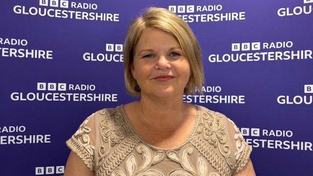 Jacqueline Gear is wearing a beige short sleeve top and has a short blonde bob. She is standing in front of a BBC Radio Gloucestershire backdrop.