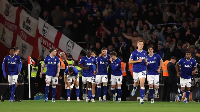 Ipswich players, wearing their blue home strip with white shorts and blue socks, celebrate as a team after scoring a goal at Southampton. The away fans can be seen celebrating behind them with Ipswich flags hanging in the stands.