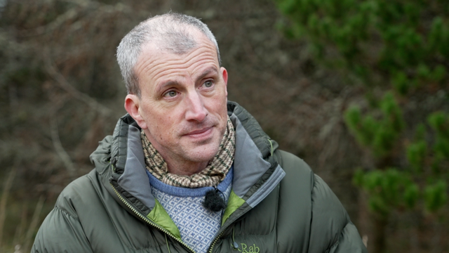 Dr Alex Langlands o Brifysgol Abertawe yn sefyll ar heneb cofrestredig ar Fynydd Cilfai, Abertawe