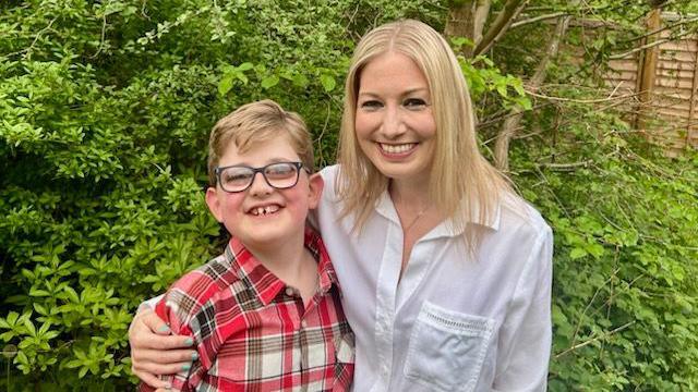 Ben and Liz Hardy, in their back garden, smiling at the camera, by a fence with green trees and bushes. Ben has on a red and whited checked shirt, glasses, and short fair hair, Liz has long blonde hair, and is wearing a white shirt.