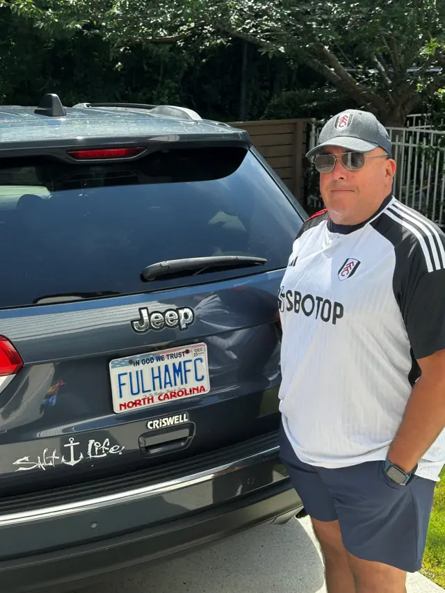 A man in a Fulham home shirt and Fulham cap stands in front of a jeep with a number plate that reads "Fulham FC"