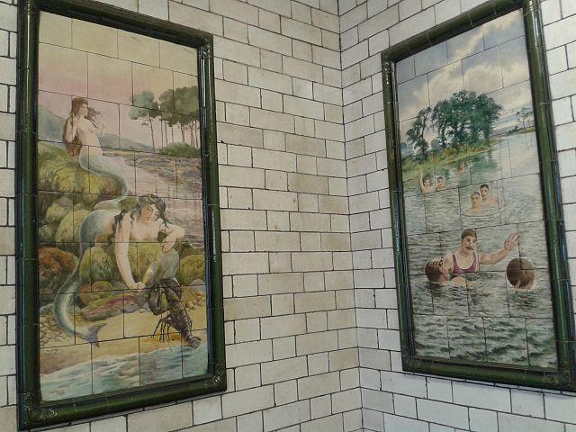 Two white-tiled walls with a picture framed on each one. The left-hand tiled image shows two mermaids, while on the left is a picture of people playing water polo. The white tiles behind them are slightly grubby.