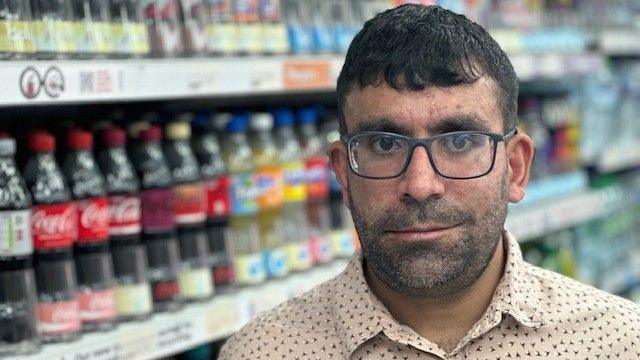 A man with dark hair and glasses wears a light-coloured shirt with a speckled pattern. He stands in front of soft drinks