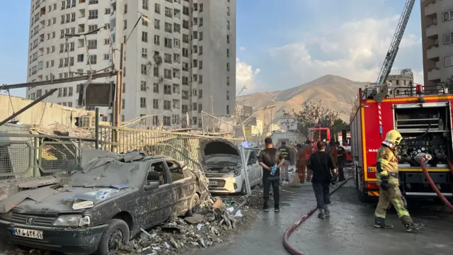 A landscape image of two cars that have been burnt out. Emergency services stand around looking at the damage and a red truck is parked on the right hand side