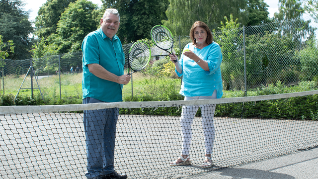 A man and a woman pose, holding tennis rackets behind a net while stood on a grey tennis court