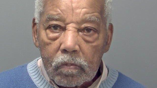 Headley's custody picture. He has white hair, eyebrows and short facial hair, and wears a white collared shirt and a blue jumper. He is looking straight ahead with a serious expression.