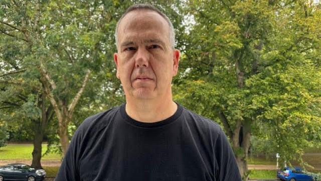 Councillor Julian Tooke stood in front of parkland with trees in Cheltenham. Some cars can be seen on a road. He has greying, short hair, and looks unimpressed. He is wearing a black t-shirt.