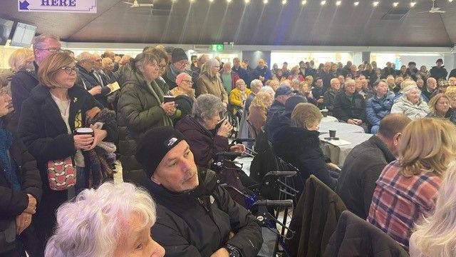 A large group of people sit in a fish and chip shop in Southport as part of a meeting to discuss a pier's future. Some people are sitting on chairs while others stand behind them. The room is full, most people are holding a cup of coffee, wearing winter coats and hats.