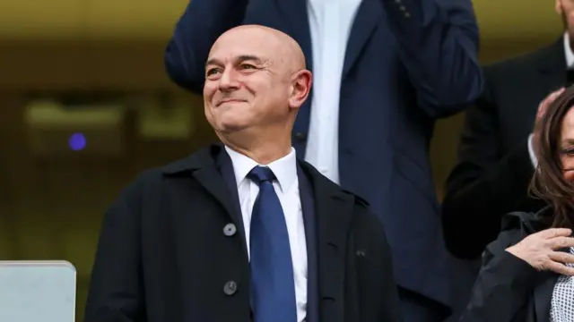 Daniel Levy smiles. He wears a shirt and tie and stands in a directors' box.
