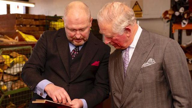 A man in a black three piece suit speaks to a man in a grey suit and purple tie. There are lots of lasts behind them. 