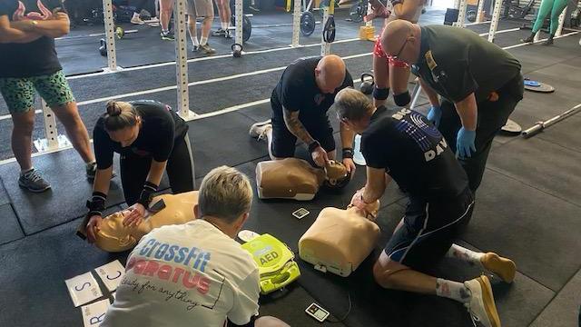 Five people kneeling down on the floor inside a CrossFit gym, practicing CPR on several dummies that are laying on black mats. There is a paramedic from Great Western Air Ambulance charity leaning over with his hands on his knees, demonstrating what to do. In the background you can see bar bells and weights on the floor.