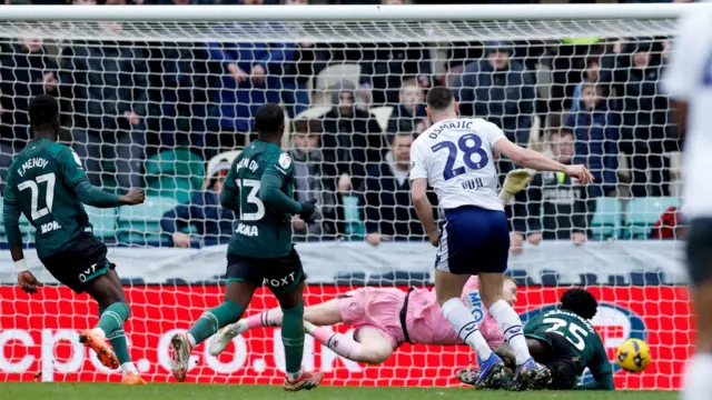 Two watford players in green face the goal running back watching as the player in white has just struck the ball into the right bottom corner of the goal as the goalkeeper in pink is at full stretch to try to reach it.