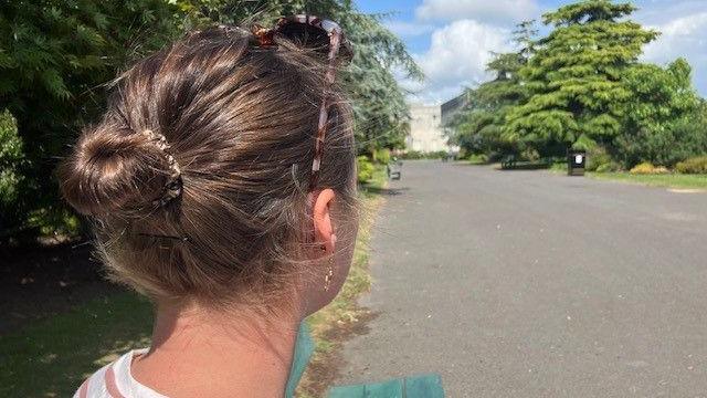 Cefn pen Louise (nid ei henw iawn) - un o'r mamau a gafodd profiadau gwael yn Ysbyty Singleton, Abertawe - yn sefyll ar bwys ffordd gyda choed ar y naill ochr