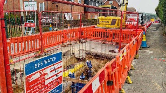 Bright orange building site barriers and red fencing enclose a large rectangular hole in thje middle of a road beside a pavement. A bright yellow digger can be seen in the stance along with many red and white traffic cones and houses in the distance on the left hand side