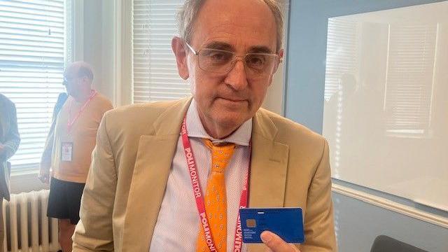 Edward Lucas, a balding middle aged man in tan suit jacket and yellow tie, holds a blue digital ID card